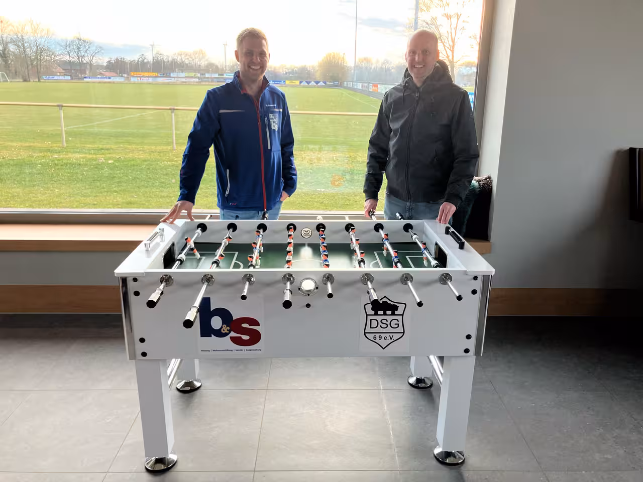 Zwei Personen neben einem weißen Tischkicker mit DSG-Logo – der Kickertisch steht im Vereinsheim mit Blick auf den Sportplatz.